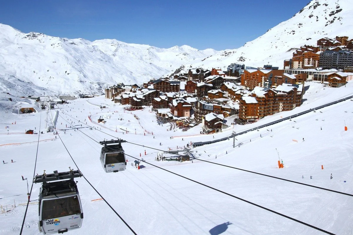 A snowy Val Thorens ski resort with a town built into the mountainside, ski lifts carrying skiers up the slope, and snow-covered buildings against a clear blue sky.