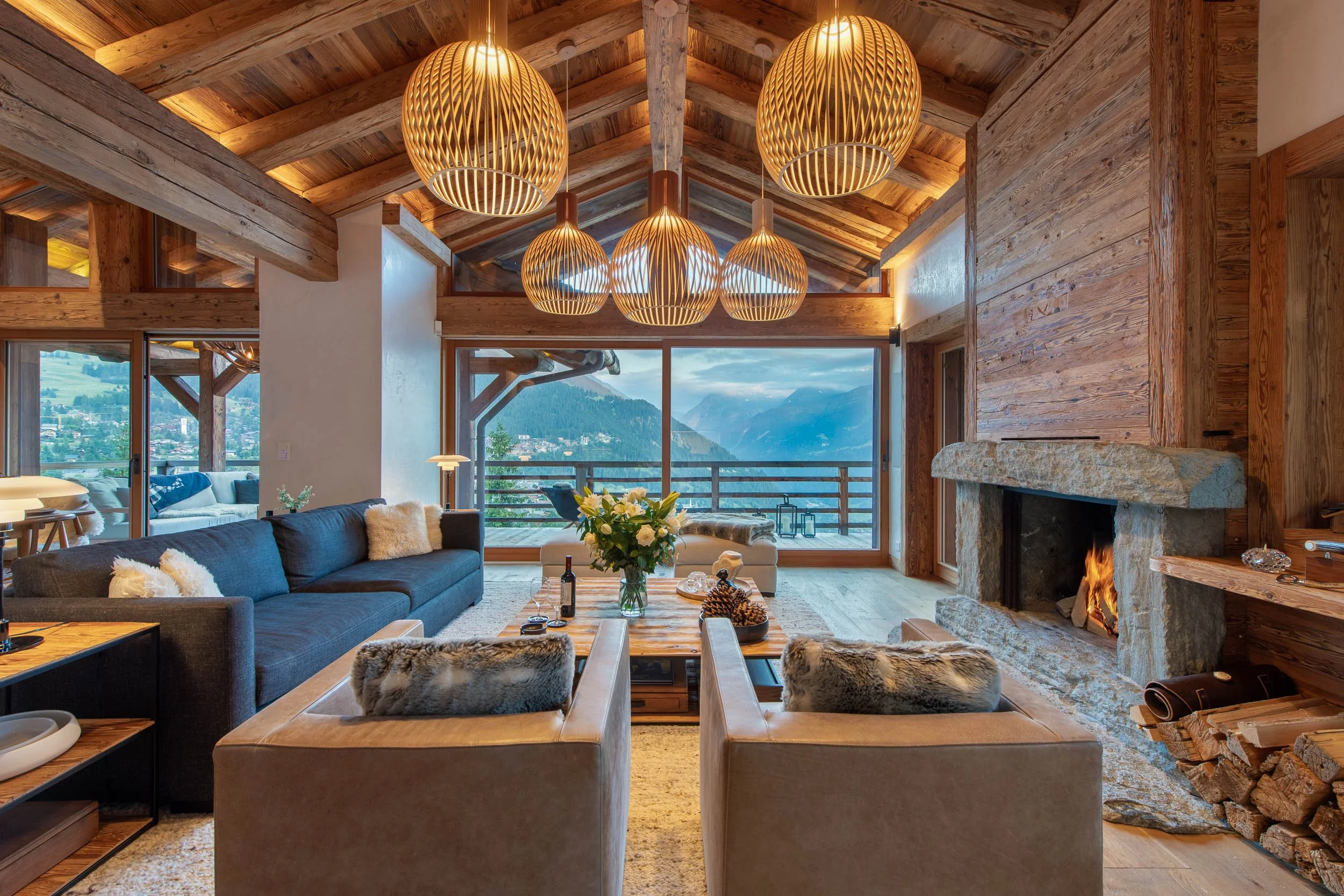 Living room with wooden beam ceiling, large glass windows showing mountain view, stone fireplace with fire, modern wooden pendant lights, dark sofa with throw pillows, coffee table with flowers and candles, and comfortable chairs with fur throws.