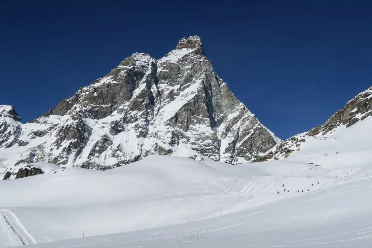 Snow-covered mountain with a rugged peak against a clear blue sky, with a group of skiers or hikers on the snow below.