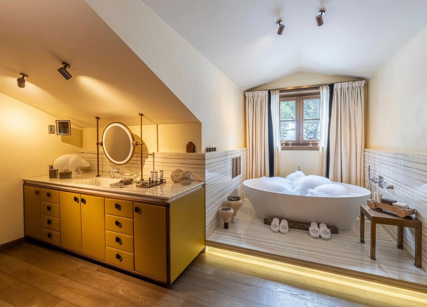 A modern bathroom with a freestanding bathtub filled with bubbles, a window with curtains, and a yellow vanity with a round mirror and various decor items.