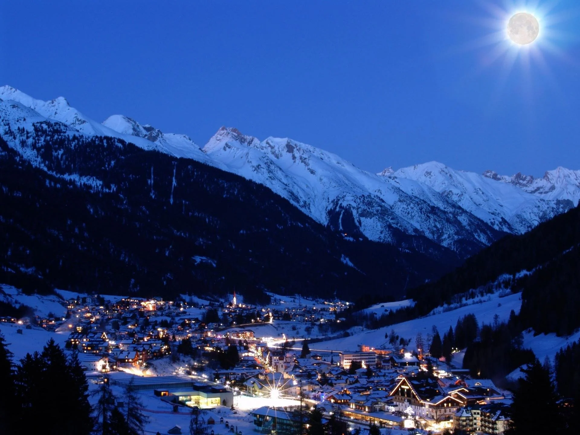 Nighttime view of a snow-covered mountain village illuminated with lights, under a bright full moon in a clear sky.