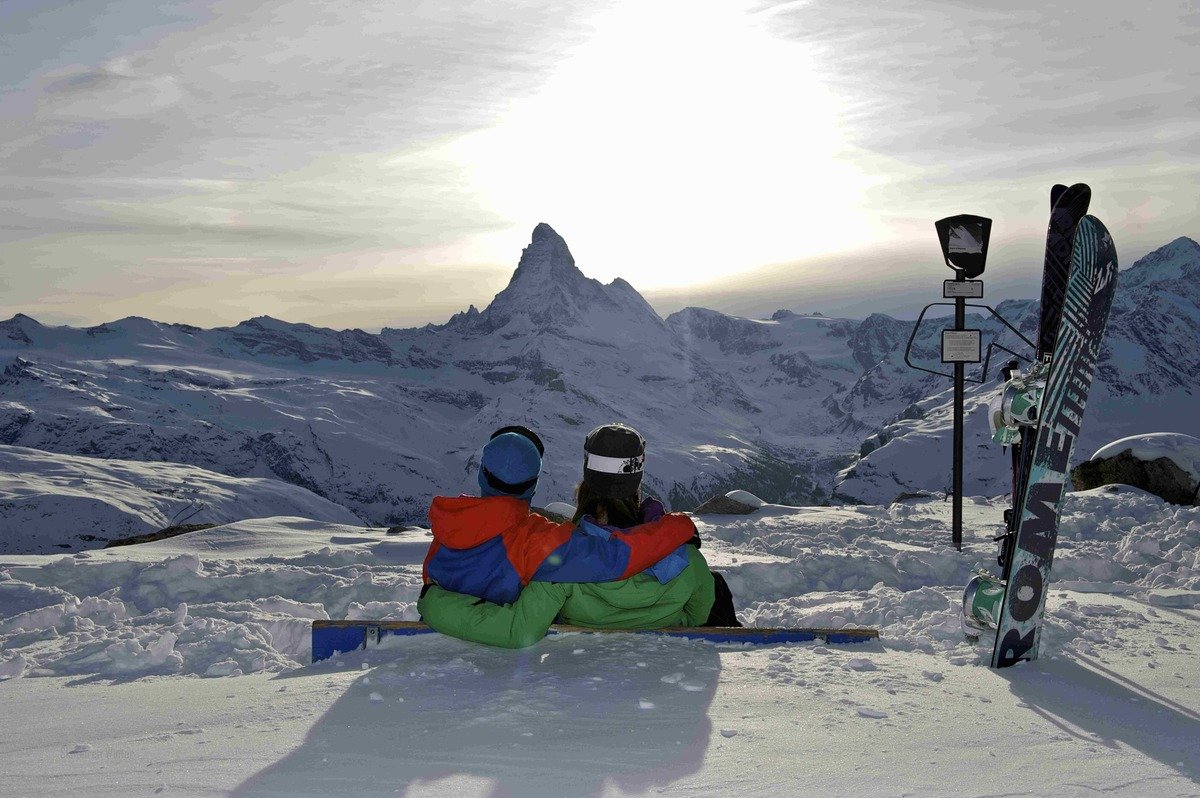 Two people sitting on the snow, one with a green jacket and the other with a multi-colored jacket, looking at the snowy Zermatt mountain landscape with the Matterhorn in the background. Ski equipment is nearby.