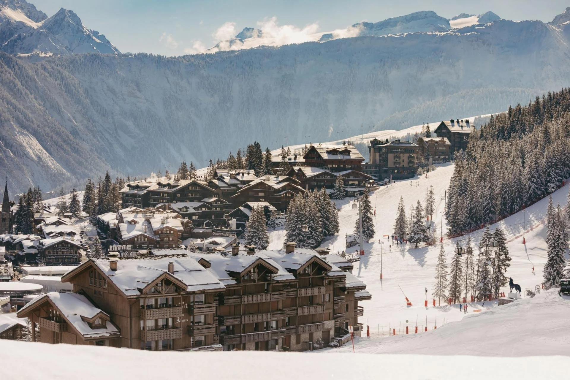 Snow-covered alpine village with chalets and hotels, surrounded by dense forest and mountains in the background, under a partly cloudy sky.