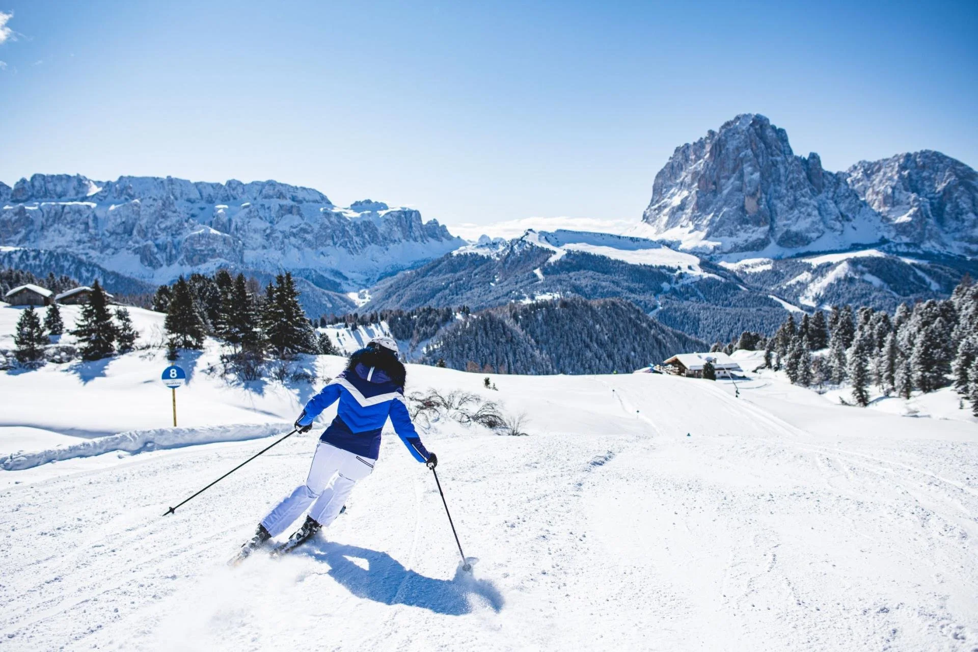 A person skiing on a snowy mountain landscape under clear blue sky with mountain peaks and pine trees in the background.
