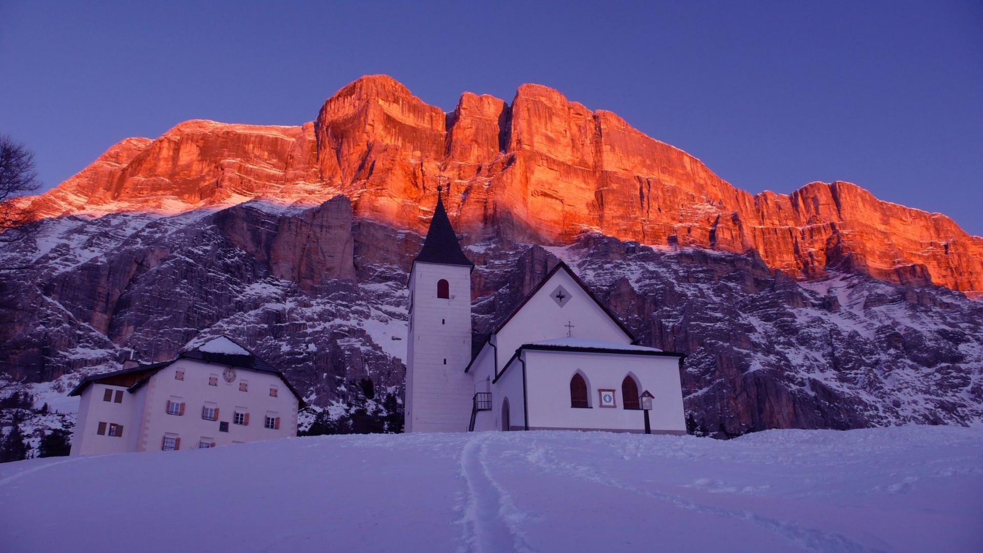 A small white church with a steeple and a building nearby, set against a mountainous backdrop with snow and ice, illuminated by the orange glow of the setting or rising sun.