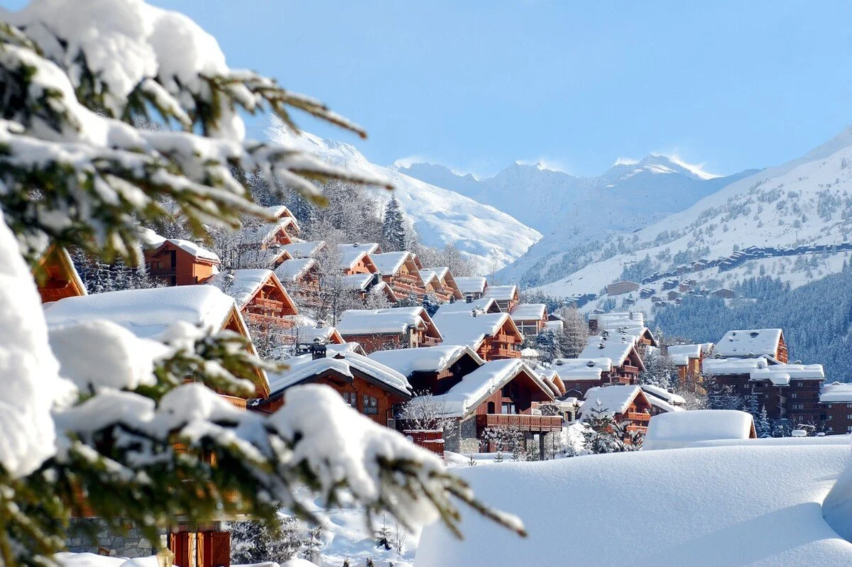 Snow-covered Meribel mountain village with wooden houses, snow-laden trees, and a clear blue sky.
