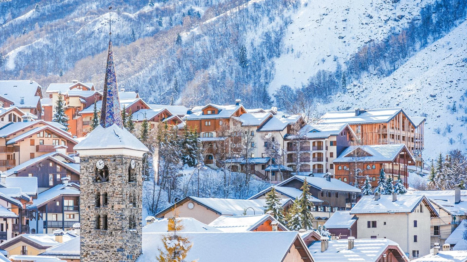 Snow-covered St Martin de Belleville village with a stone clock tower and various chalets and buildings on a mountainside during winter.