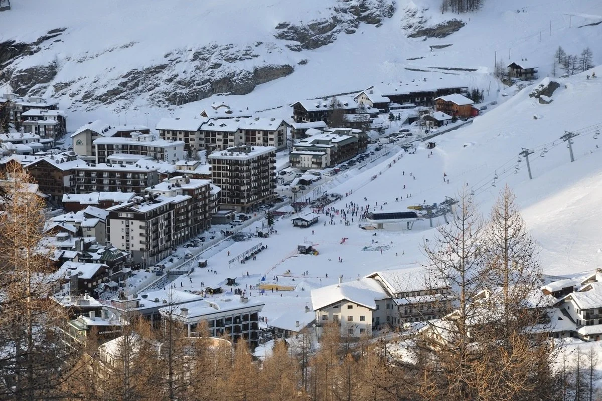 A snowy mountain town with ski slopes and buildings covered in snow, with skiers on the slopes and chairlifts in the background.