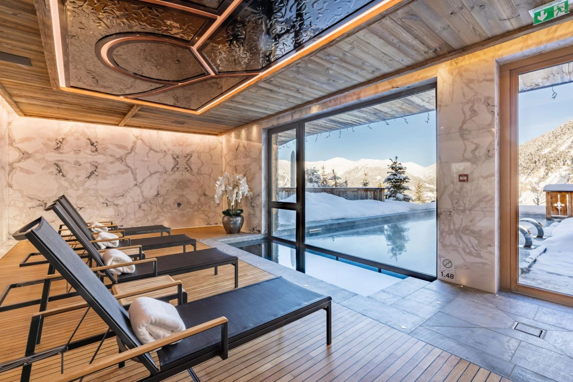Indoor sauna or relaxation room with wooden floors and walls, black lounge chairs with rolled white towels, large window showing snowy mountains and snow-covered outdoor area, and ceiling with decorative black and wood pattern.