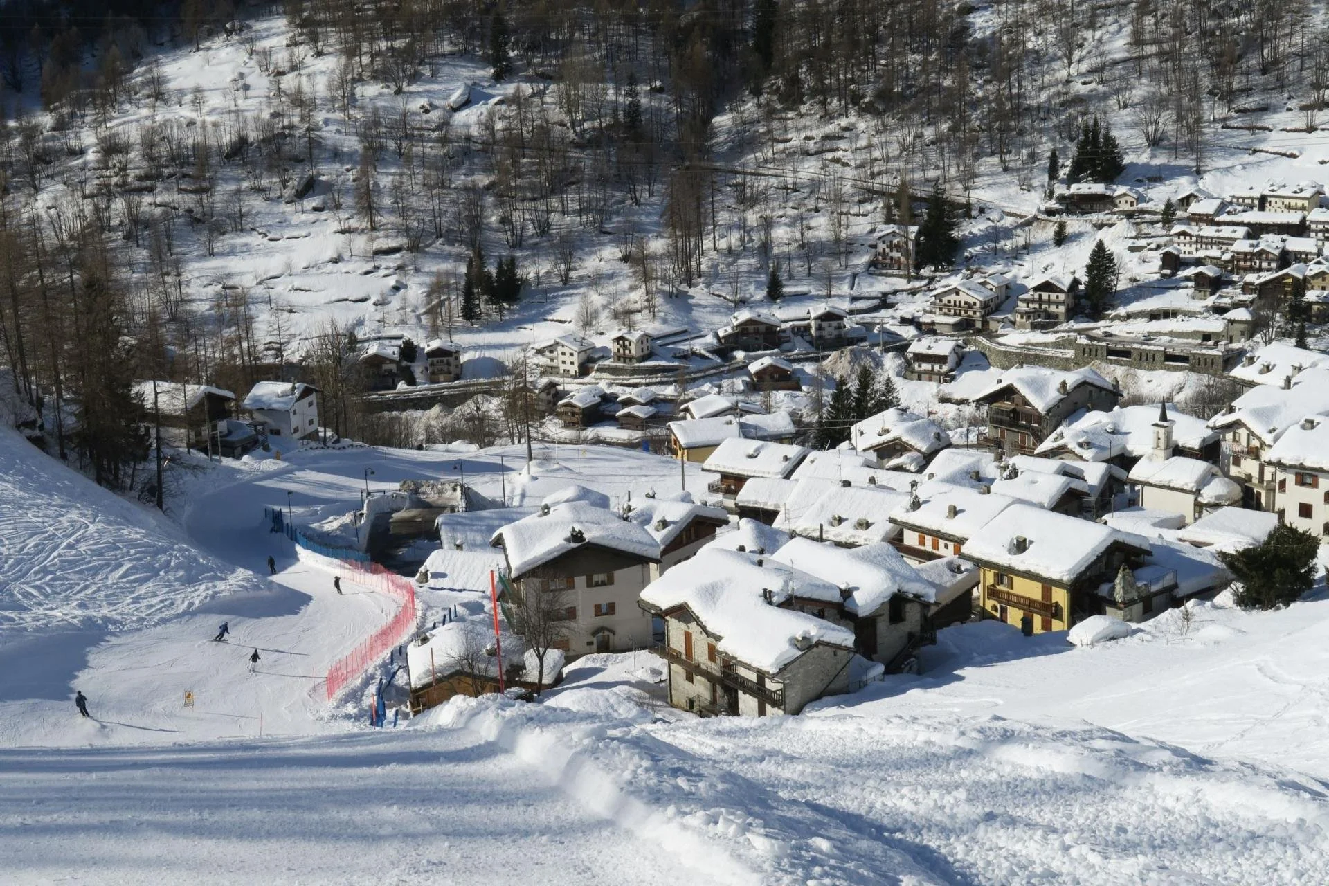 Snow-covered ski resort with skiers, ski lift, and chalet-style buildings in a mountainous landscape.
