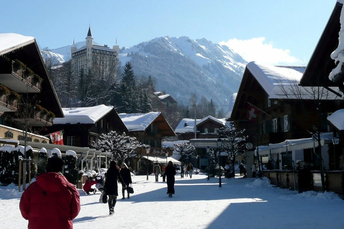Snow-covered Gstaad village street with chalet-style buildings, people walking, and a mountain in the background, during winter.