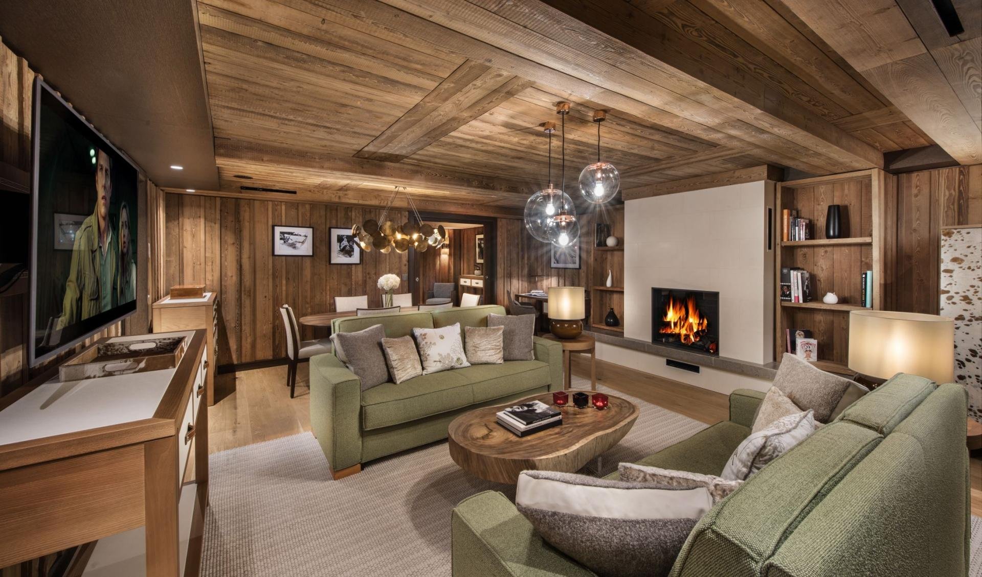A cozy living room with wooden ceiling and walls, featuring a fireplace, green sofas, a wooden coffee table, a flat-screen TV, and various decorative items and artwork.