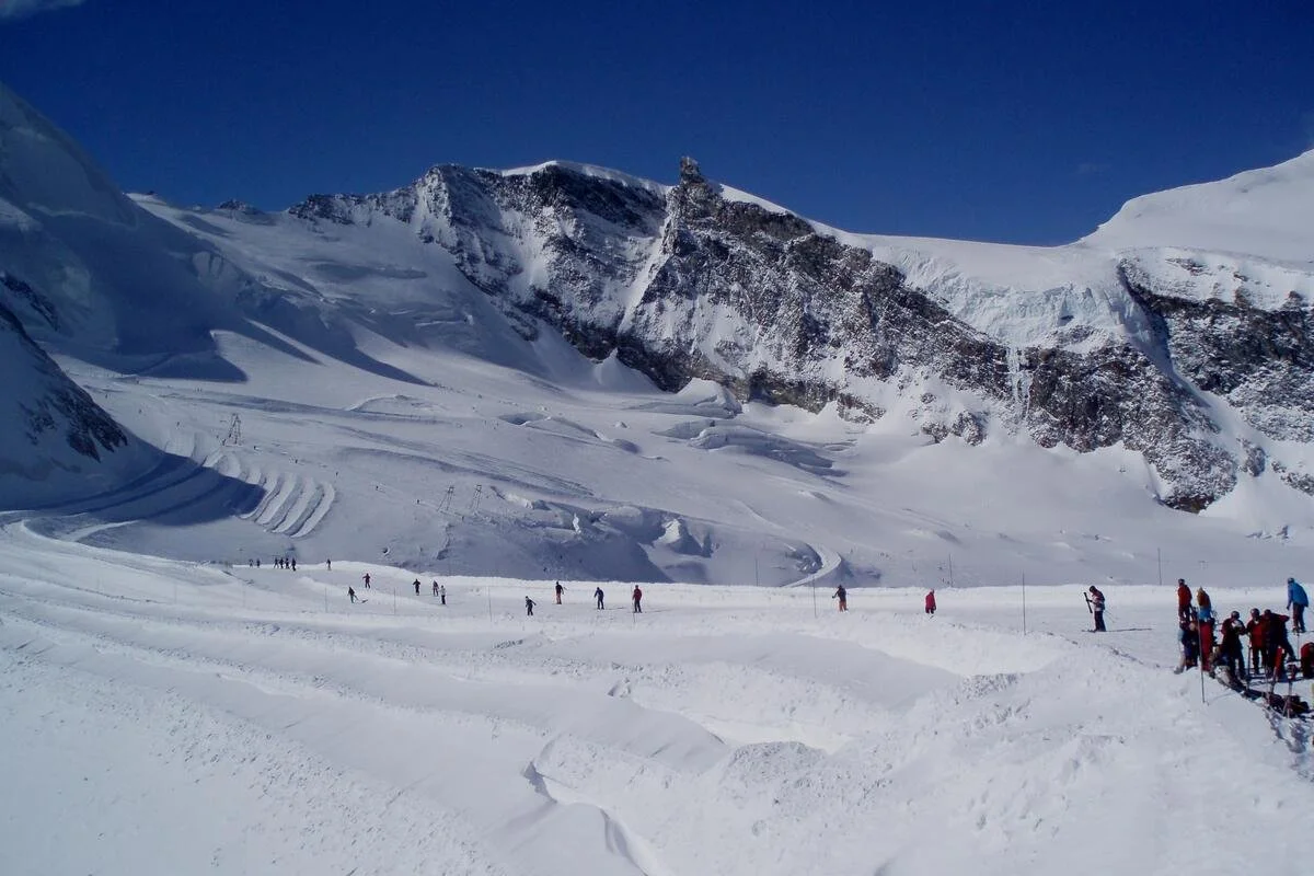 Snow-covered Saas-Fee mountain landscape with skiers and snowboarders, clear blue sky, and rugged peaks.