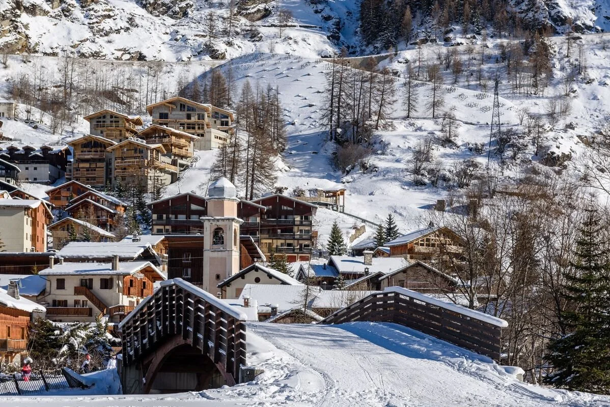 Snow-covered Tignes village with chalets and a small arched bridge in the foreground, set against a snowy mountain backdrop with trees.
