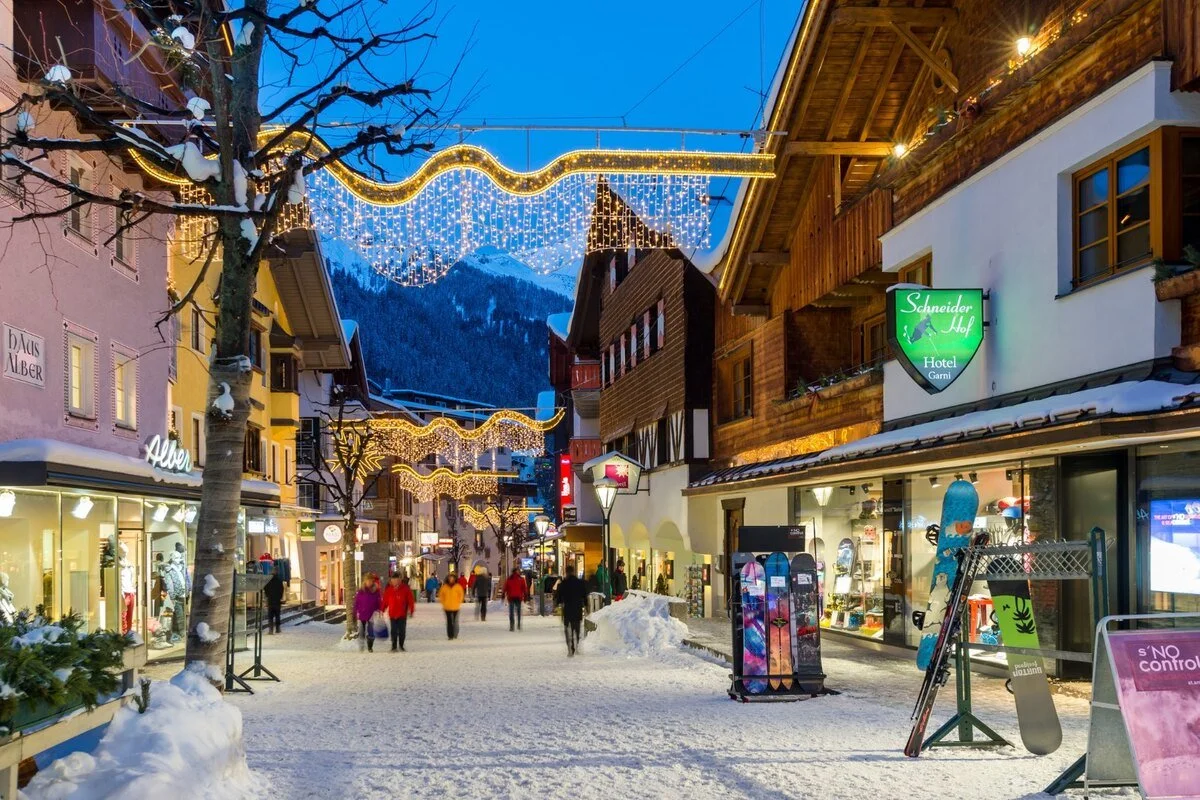 A lively winter street scene in a European-style mountain town during evening with snow-covered ground, outdoor shops, holiday lights, and pedestrians wearing winter clothing.