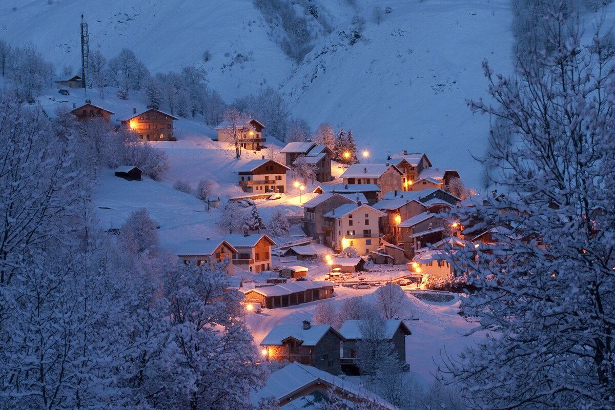 A snowy St Martin de Belleville village at dusk with houses illuminated by warm yellow lights.