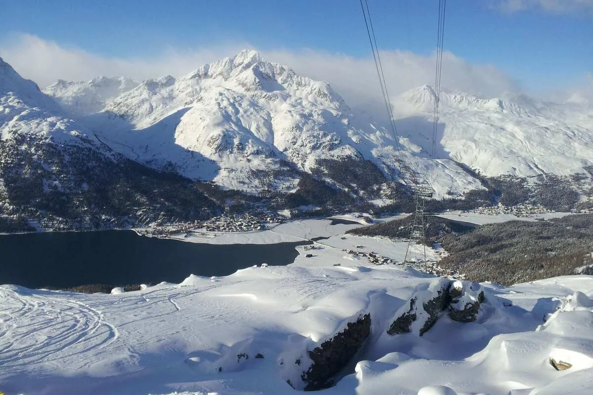 Snow-covered St Moritz mountains with St Moritz lake and small town below, with ski tracks and a ski lift cable in the foreground.