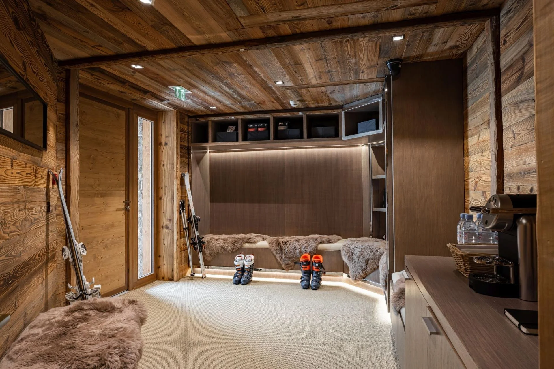 A cozy indoor space with wooden walls and ceiling, featuring skis and ski boots, and a sitting area with faux fur blankets and water bottles.