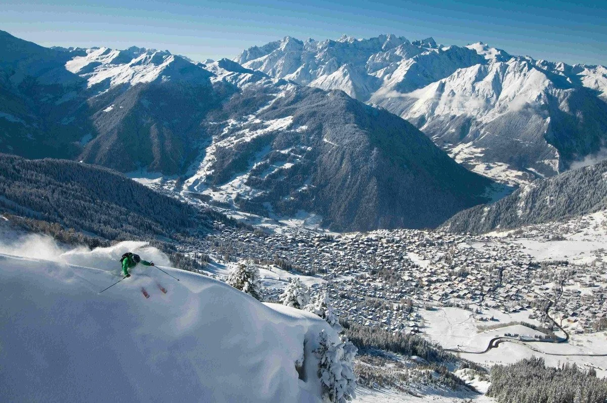 A skier in green gear skiing down a snowy mountain slope in Verbier in the foreground, with a mountain town below and snow-covered peaks in the background.
