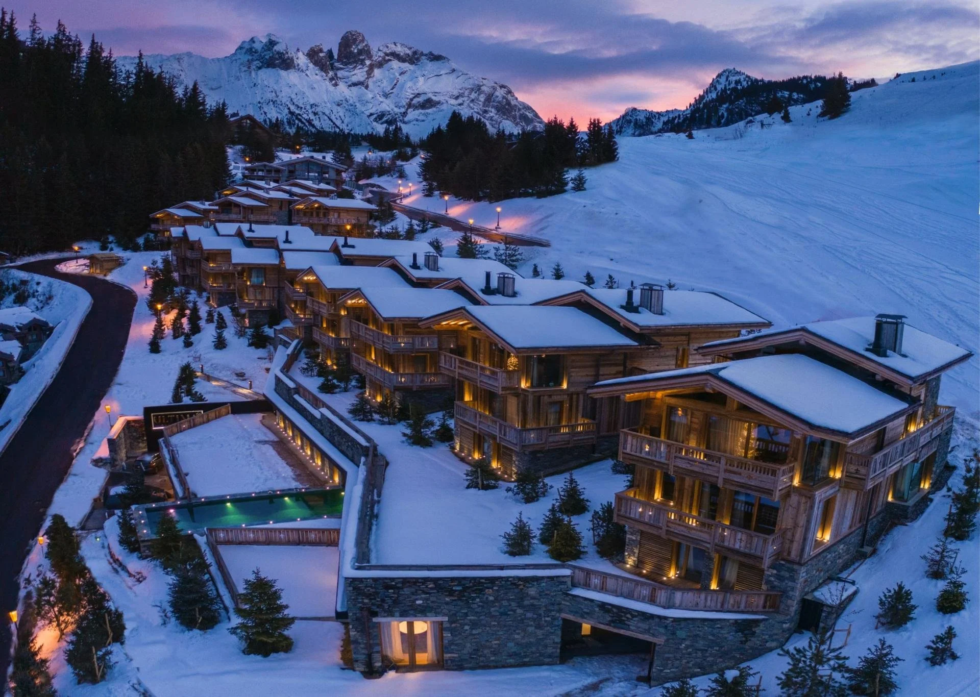 Aerial view of a snow-covered mountain landscape with a ski slope in the background and a cluster of illuminated wooden lodge-style buildings in the foreground during dusk.
