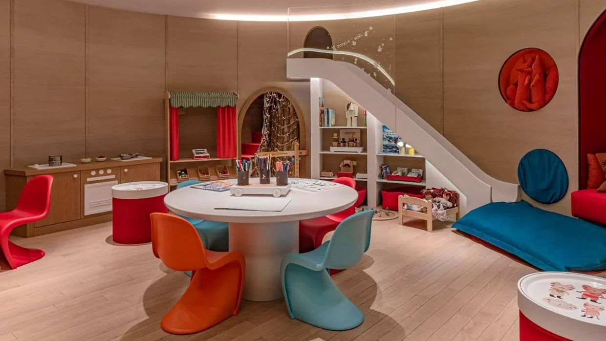 Colorful children's playroom with a round table, chairs, bookshelf, stairs, and soft seating area.