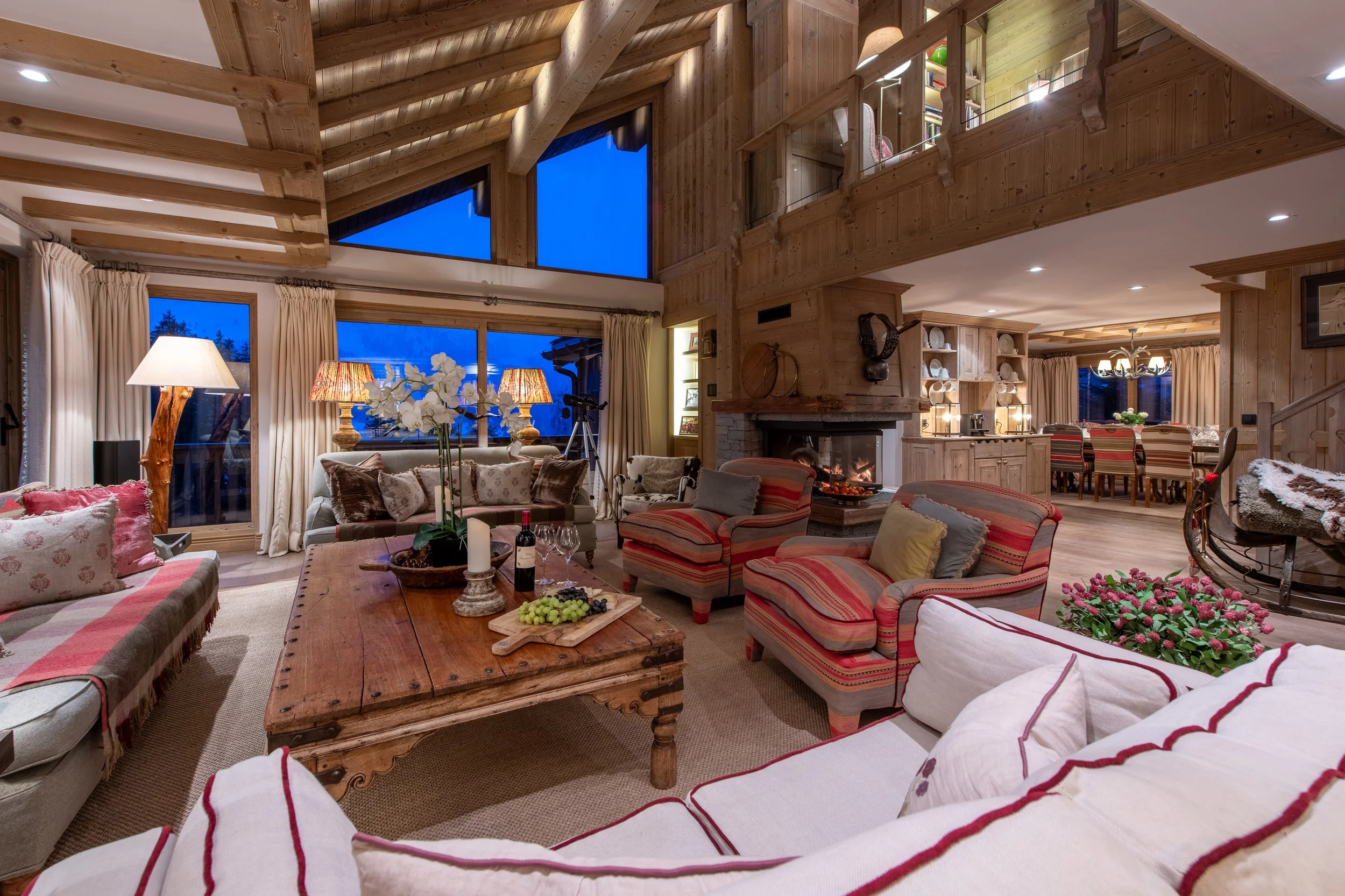 A spacious living room with large windows showing a night sky outside. The room features wooden beams and paneling, with comfortable sofas, a large wooden coffee table with grapes and candles, and a fireplace. There are decorative lamps, plants, and curtains, with a dining area visible in the background.