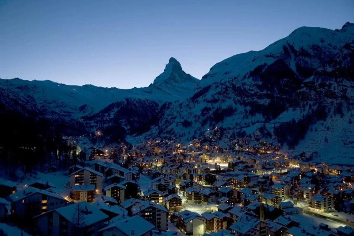 Snow-covered Zermatt mountain town at dusk with illuminated buildings and the Matterhorn mountain in the background.