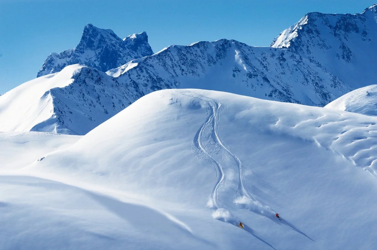 Two skiers skiing down a snow-covered mountain in Chamonix with mountain peaks in the background.