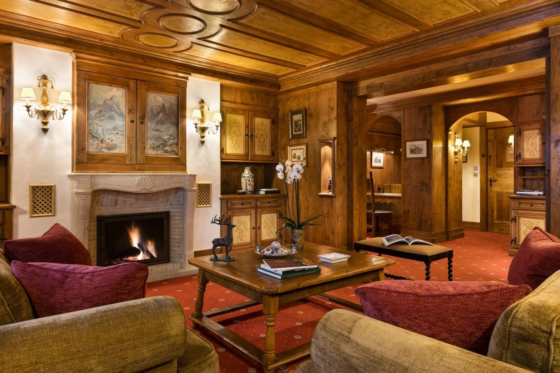 Cozy living room with wooden paneled walls and ceiling, a fireplace with a stone mantel, comfortable chairs, a coffee table with books, decorative items, and framed artwork on the walls, illuminated by wall sconces.