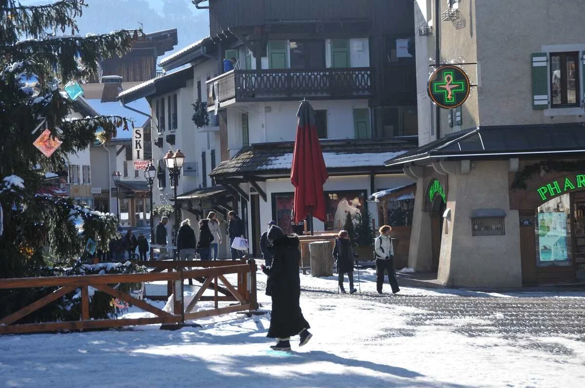 A snowy street scene in Megeve with shops, people walking, and a pharmacy with a green cross sign.