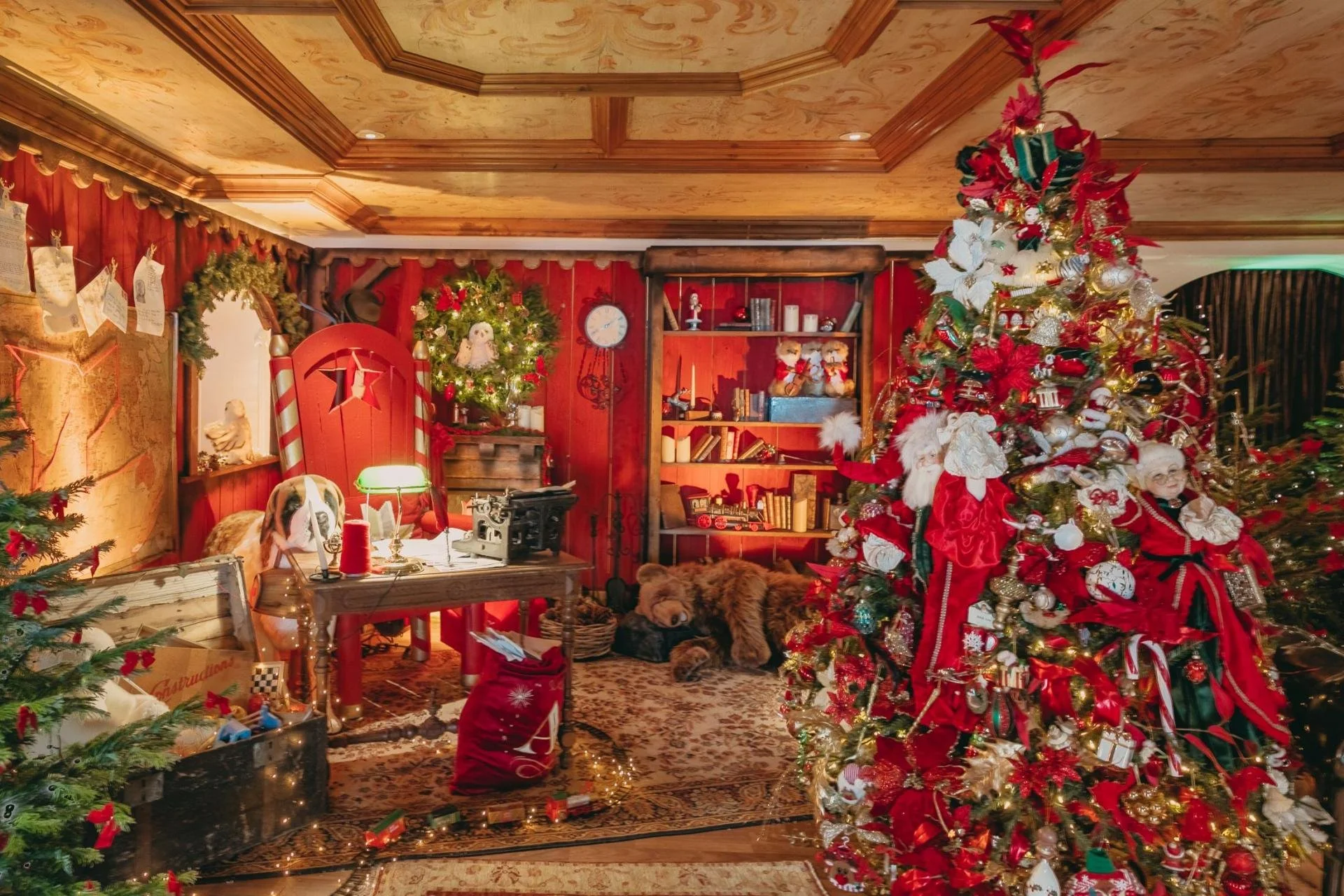 A decorated Christmas room with a large Christmas tree adorned with ornaments, ribbons, and lights. The room features red walls, a bookshelf with holiday decorations, and a festive desk with a lamp and sewing machine. There are holiday figures, stuff