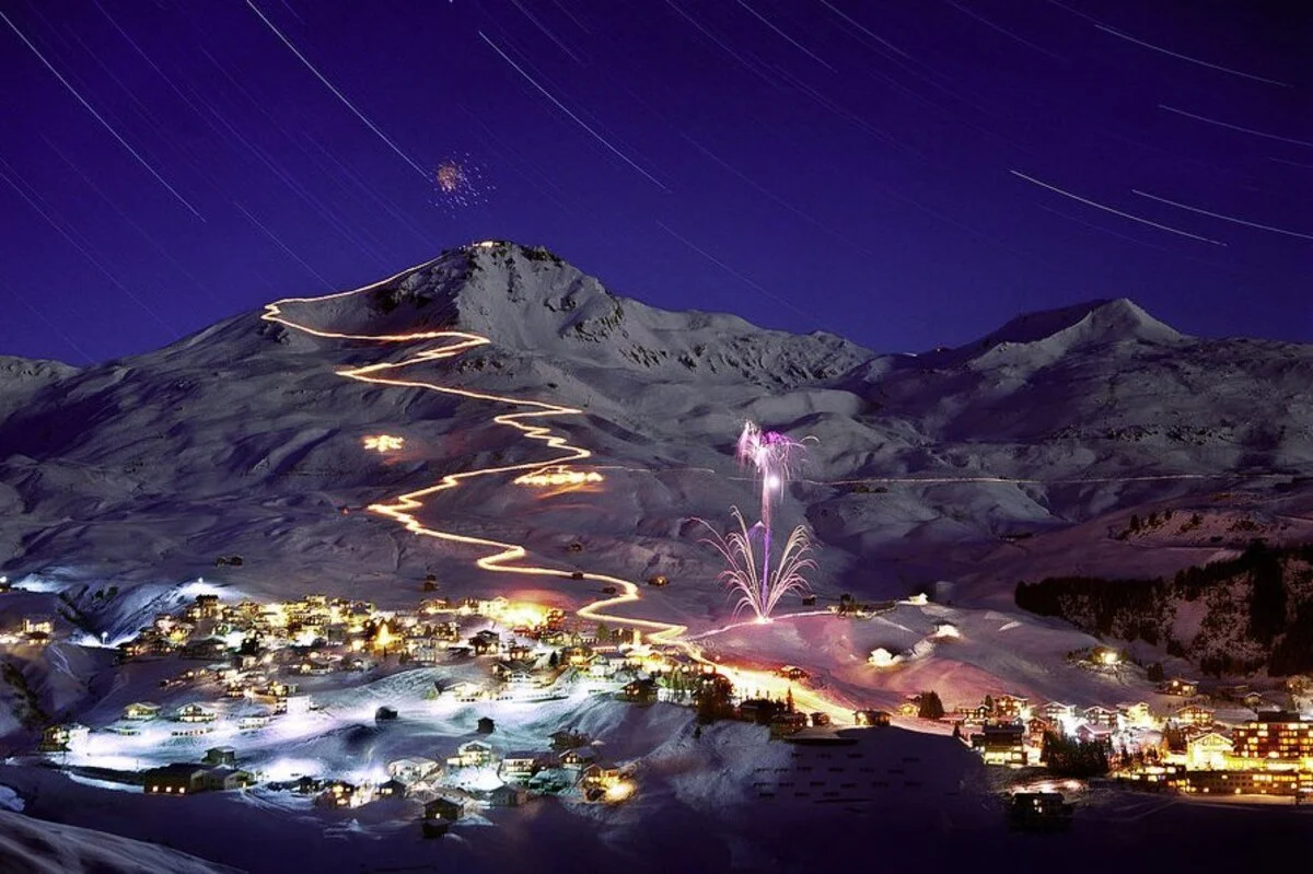Snow-covered Arosa mountain at night with star trails in the sky and fireworks near a village at the base, with a winding lit trail leading up the mountain.