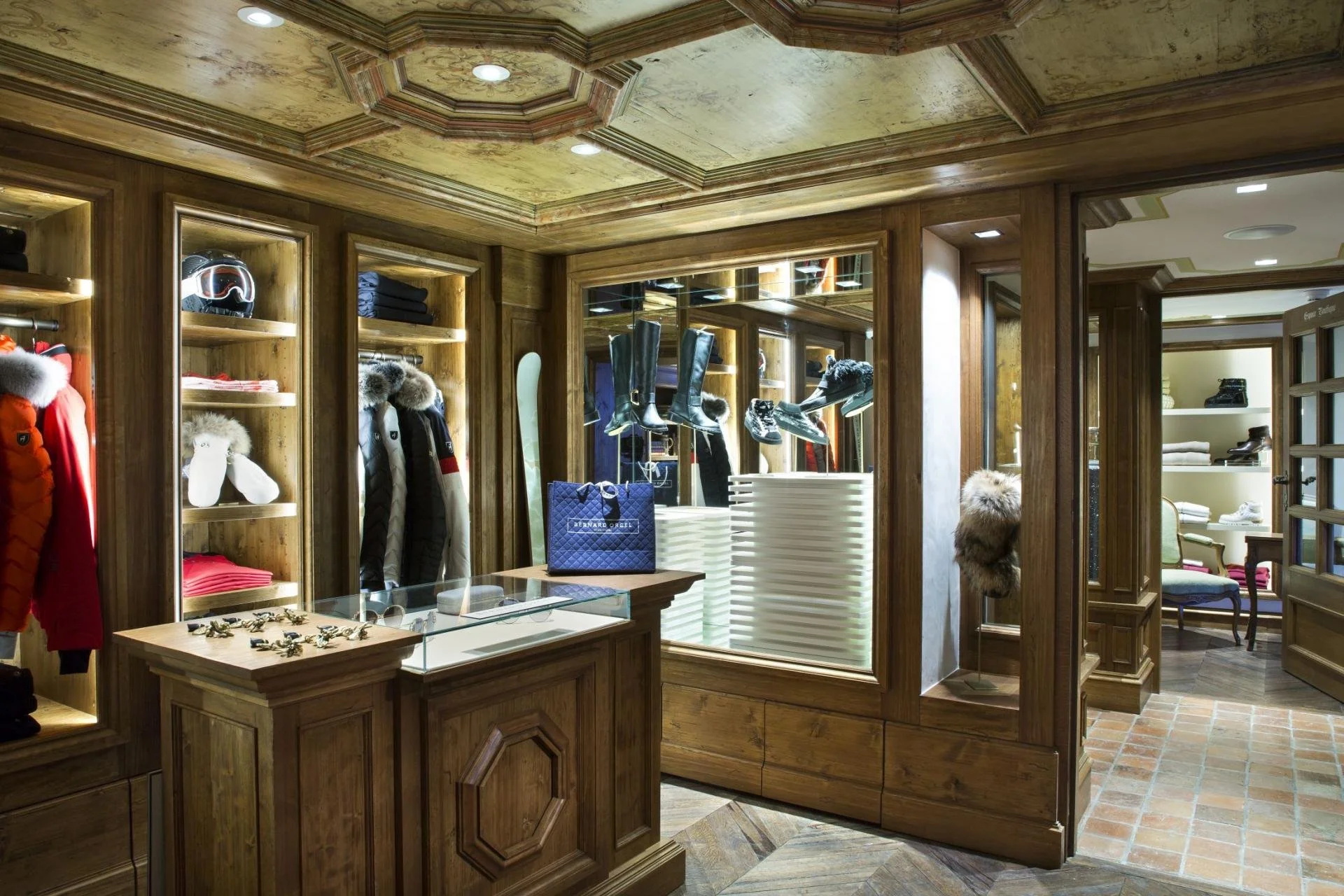Interior of a clothing store with wooden shelves and display cases, displaying winter jackets, hats, and accessories, with reflective glass and warm lighting.