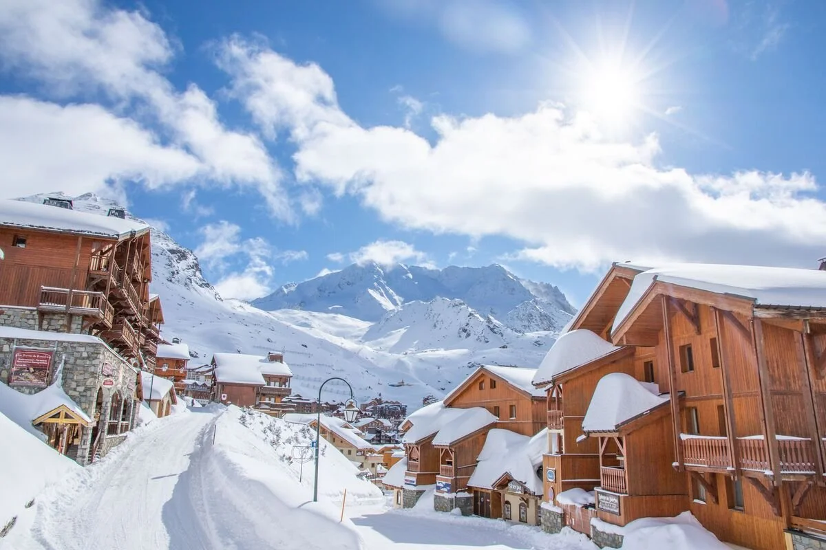 Snow-covered chalet-style buildings in a Val Thorens under a bright blue sky with clouds and sunlight.