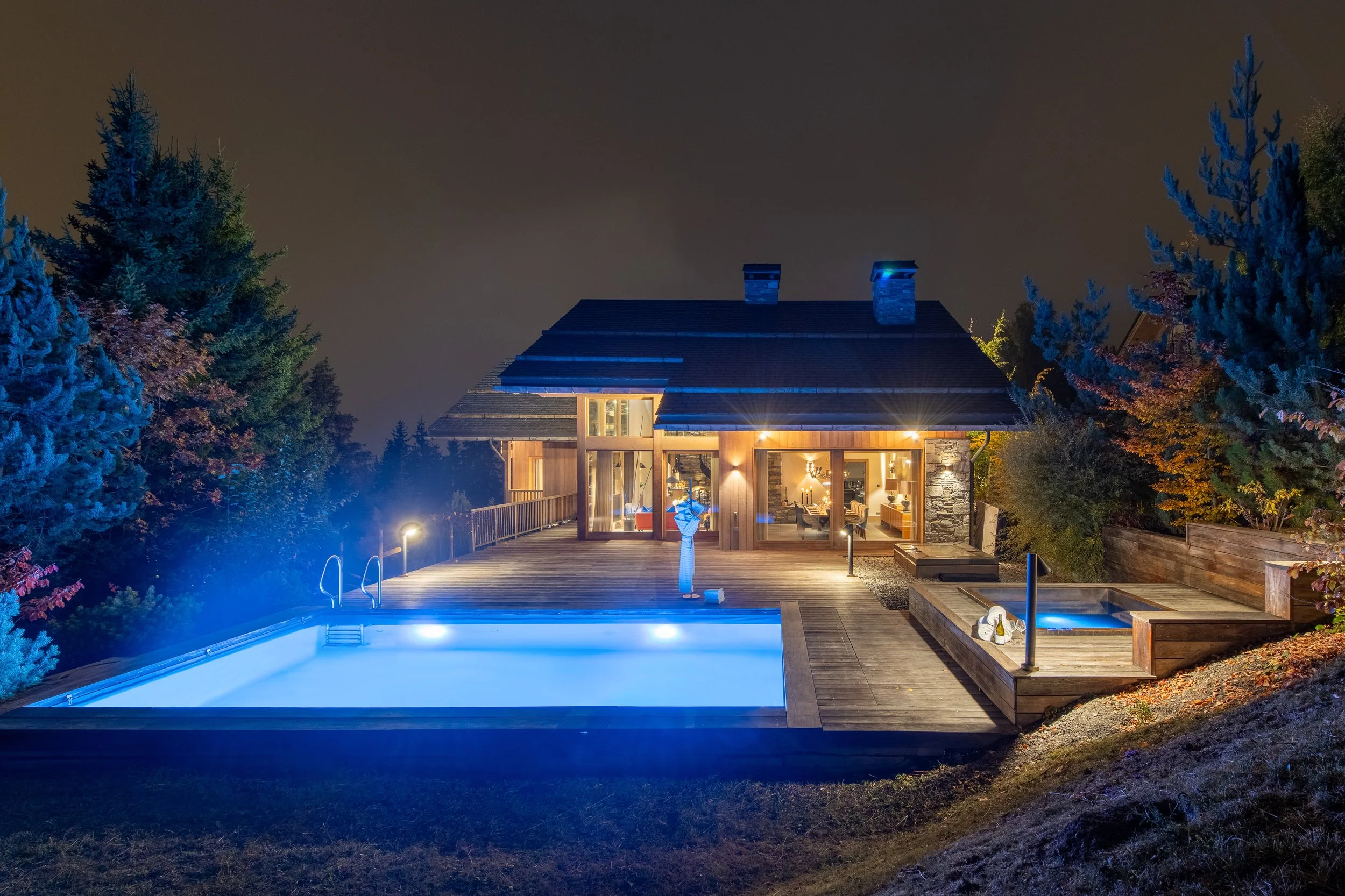 Night view of a modern house with large glass windows, a swimming pool, and a deck, surrounded by trees with some colored autumn leaves, illuminated by outdoor lighting.