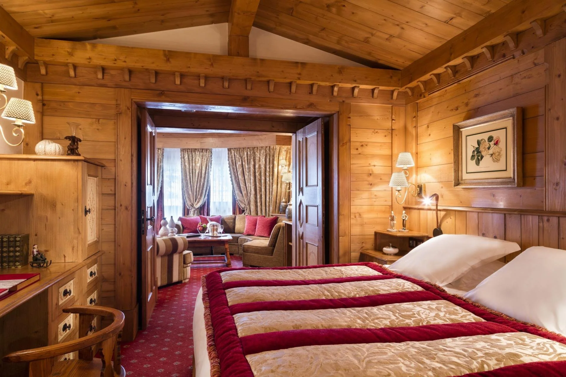 Hotel room with wooden walls and ceiling, two beds with red and gold bedding, bedside lamps, and framed floral artwork. An open doorway leads to a sitting area with curved sofa, armchair, and large windows with curtains.