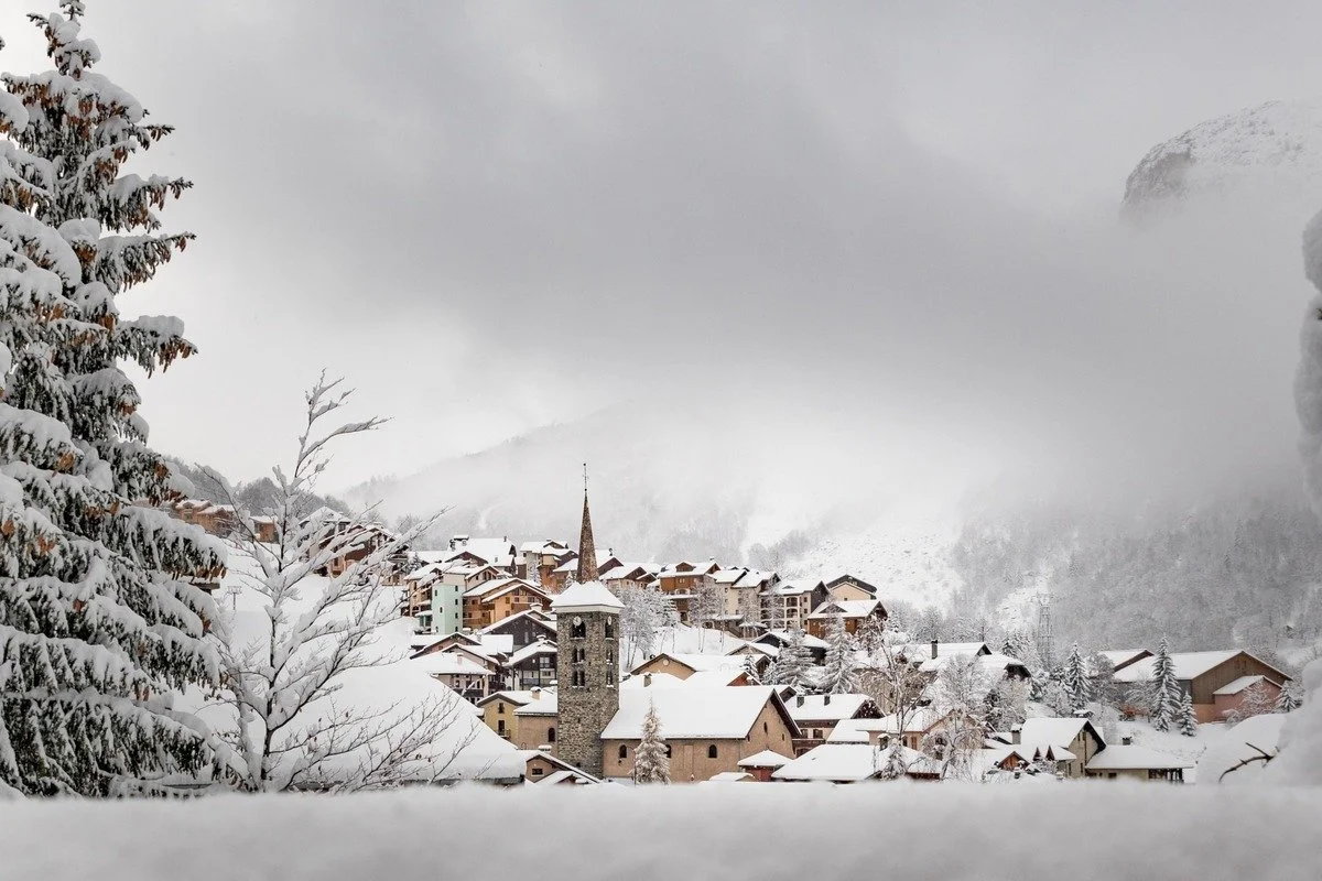 Snow-covered St Martin de Belleville village with church steeple, surrounded by snow-laden trees and mountains in the background on a cloudy winter day.