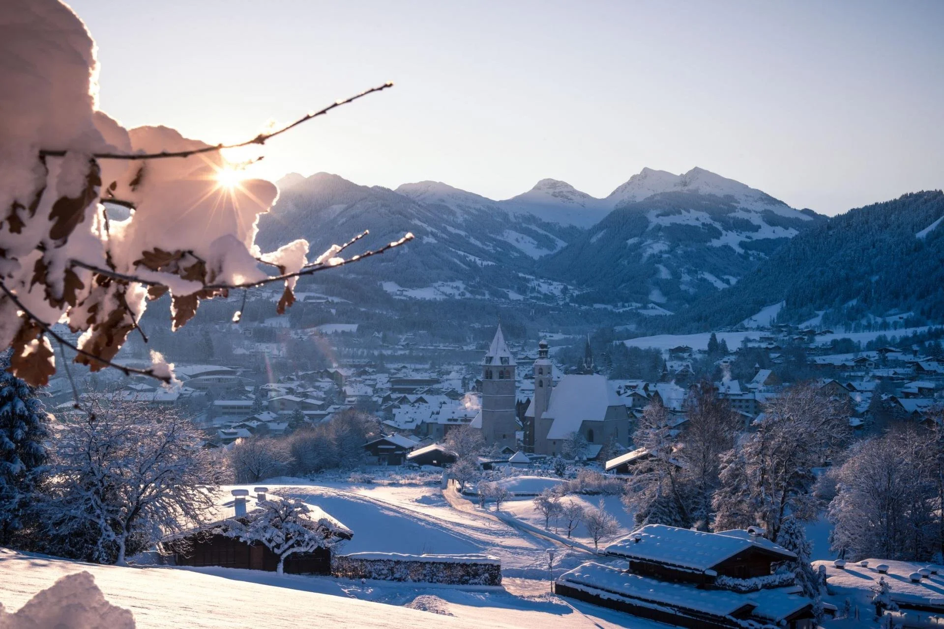 Snow-covered Kitzbuhel village in the mountains during sunrise, with snow-laden trees and a church in the center.