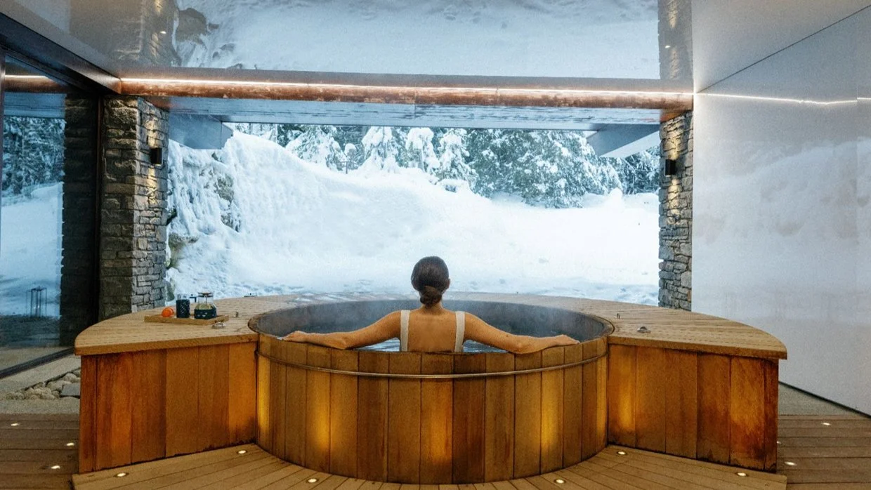 Woman relaxing in a wooden hot tub indoors, with a large window showing a snowy landscape outside.