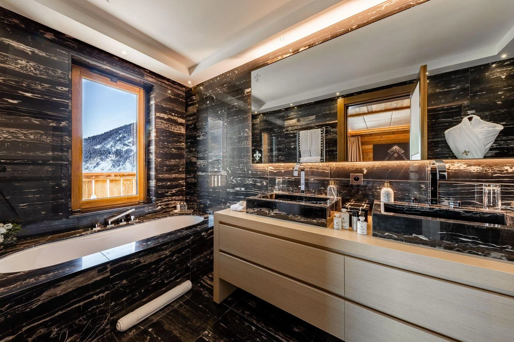 Luxurious bathroom with black marble walls and counters, a large mirror, a window showing a snowy mountain landscape, a bathtub, and a sink with toiletries on a beige wooden vanity.