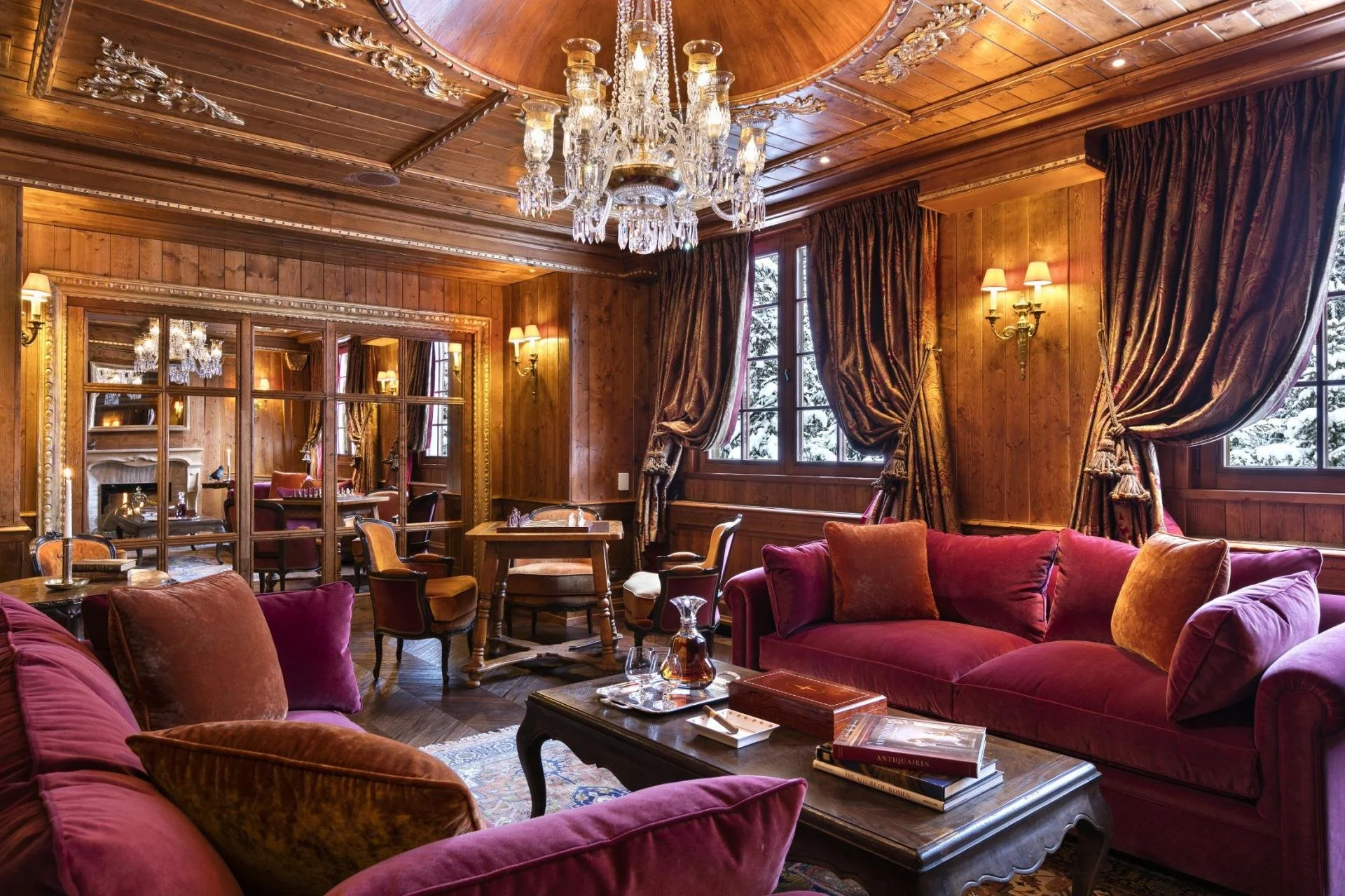 Elegant room with wood-paneled walls, large windows with heavy curtains, crystal chandelier, and plush red velvet sofas and armchairs.