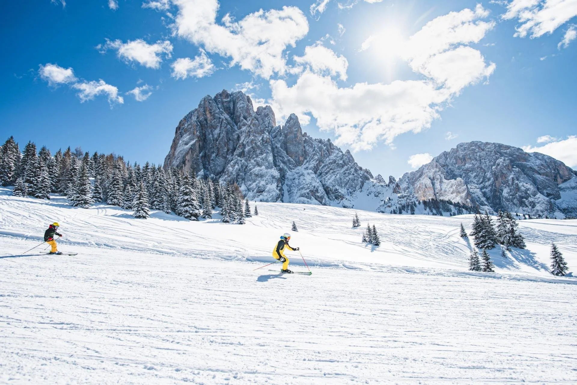 Two skiers on a snowy mountain slope with a backdrop of rocky peaks, pine trees, and a partly cloudy blue sky.