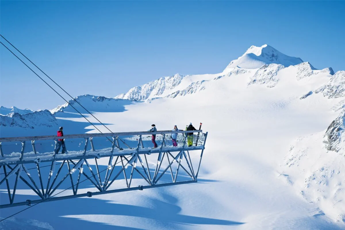 Group of four people standing on an observation platform in Solden, with a snow-covered mountain in the background and clear blue sky.