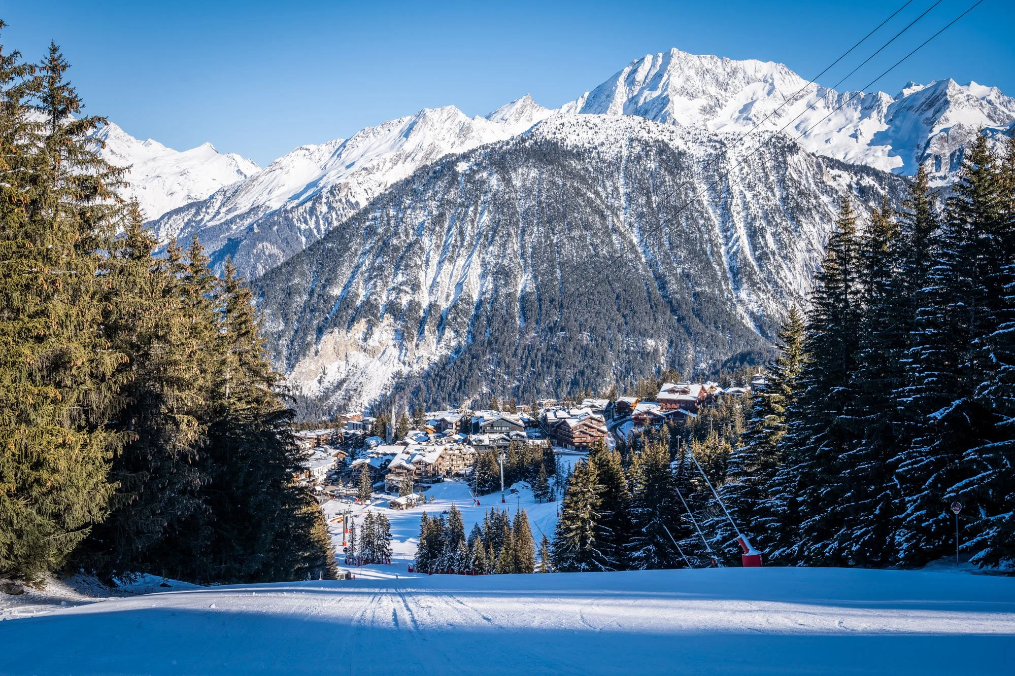 Snow-covered Courchevel 1850 ski slope with trees on both sides leading to a small village at the base of snow-capped mountains under a clear blue sky.