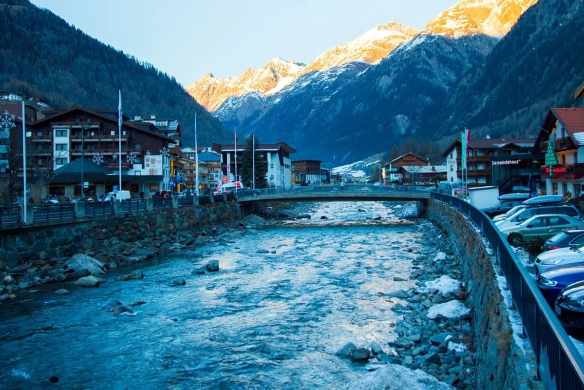 Solden mountain town with snow-covered peaks in the background, a river running through the center, and buildings and parked cars along the riverbank.