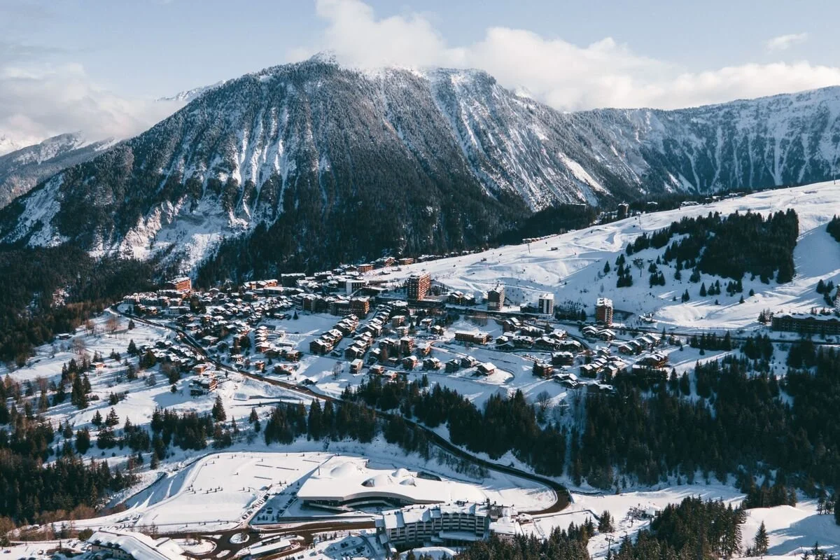 A snowy Courchevel 1650 mountain village nestled in a valley with tall snow-covered peaks in the background and a winding road leading into the town.