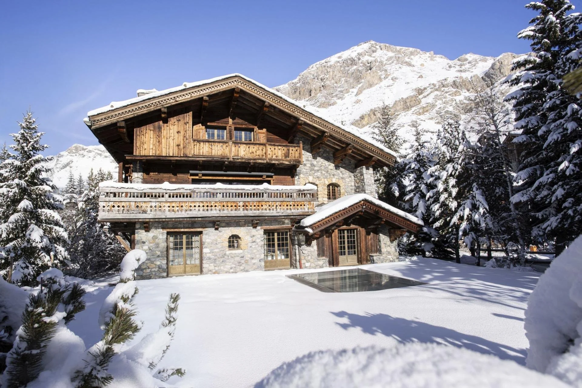 A rustic wooden and stone house amid snow-covered trees and ground, with snow-capped mountains in the background under a clear blue sky.