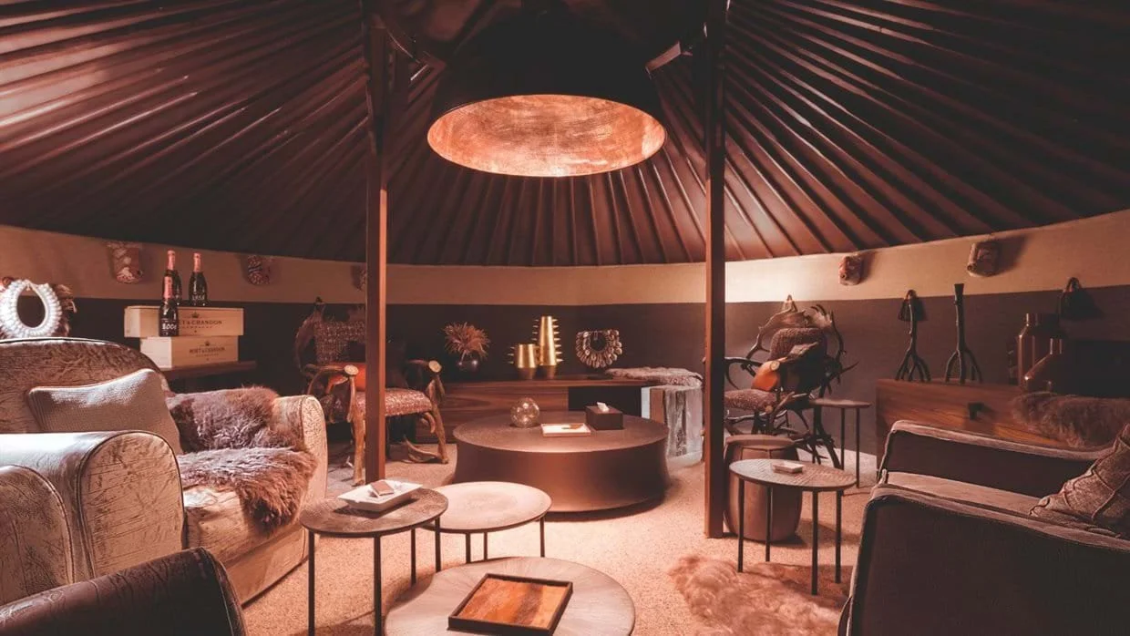 Luxurious, cozy lounge area with plush sofas, patterned chairs, and small side tables. Warm, dim lighting from a central hanging fixture creates an intimate atmosphere, with decorative items like vases and framed art on the walls.