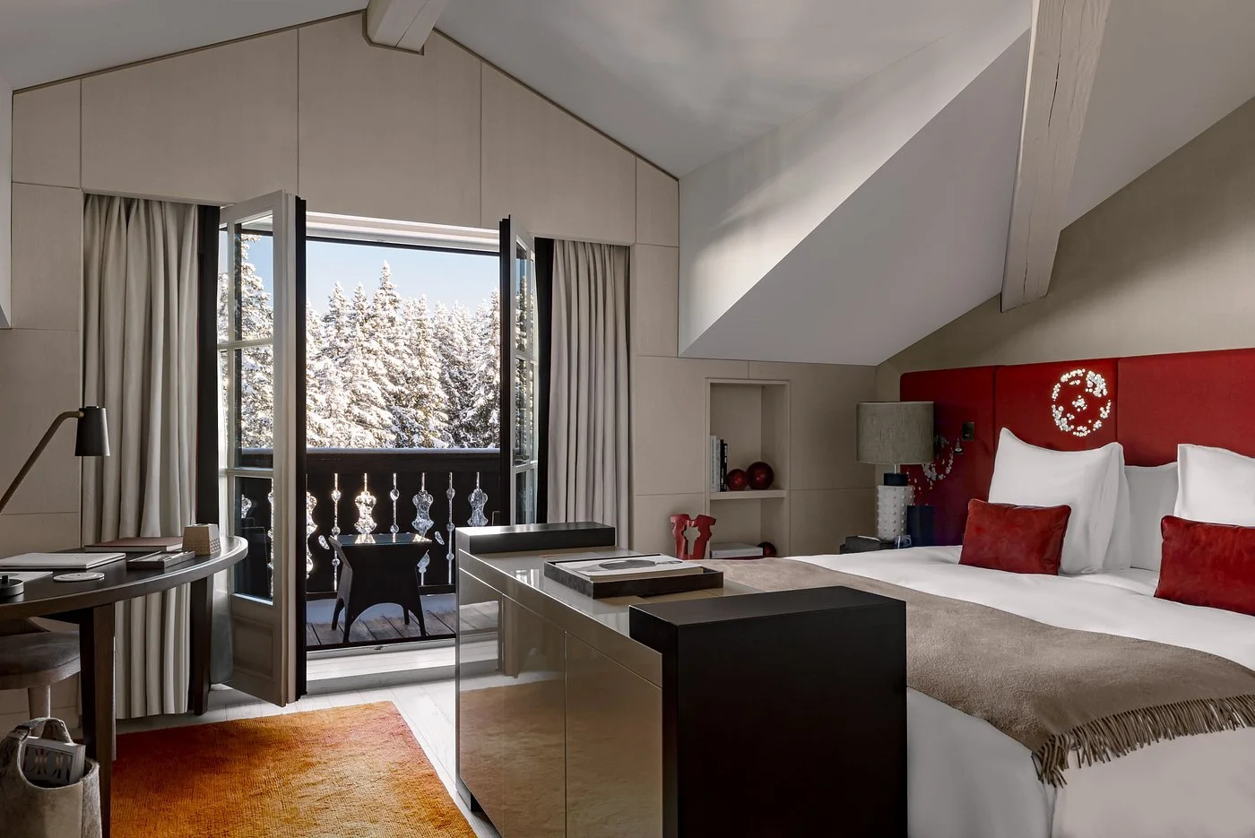 Hotel room with a balcony view of snowy trees, a bed with white sheets, red pillows, and a red headboard, a small black desk, and a beige rug.