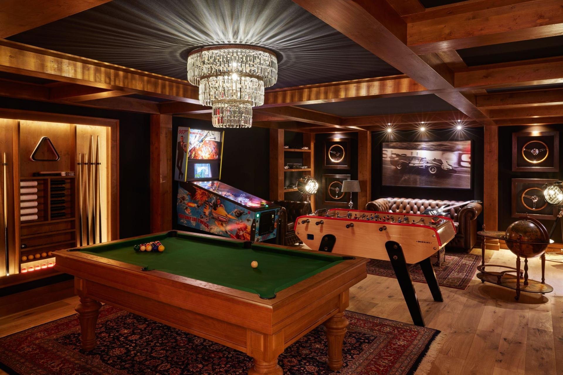 A spacious luxury game room with wooden walls and ceiling, featuring a crystal chandelier, pool table, pinball machine, foosball table, large television, and various wall art and decor.